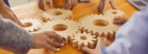 Mains de plusieurs personnes emboîtant des engrenages en bois sur une table, symbolisant l’organisation de la comptabilité associative.
