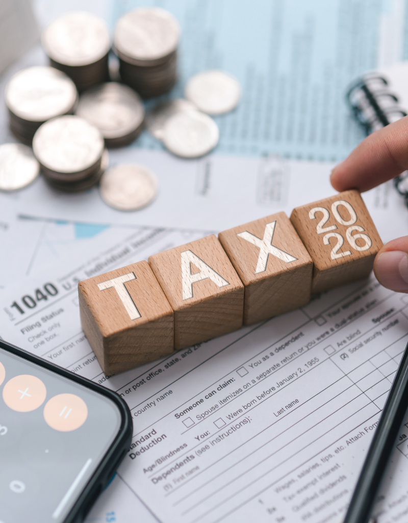 Blocs en bois formant les mots « TAX 2026 » sur des documents fiscaux, avec une calculatrice et des pièces de monnaie.